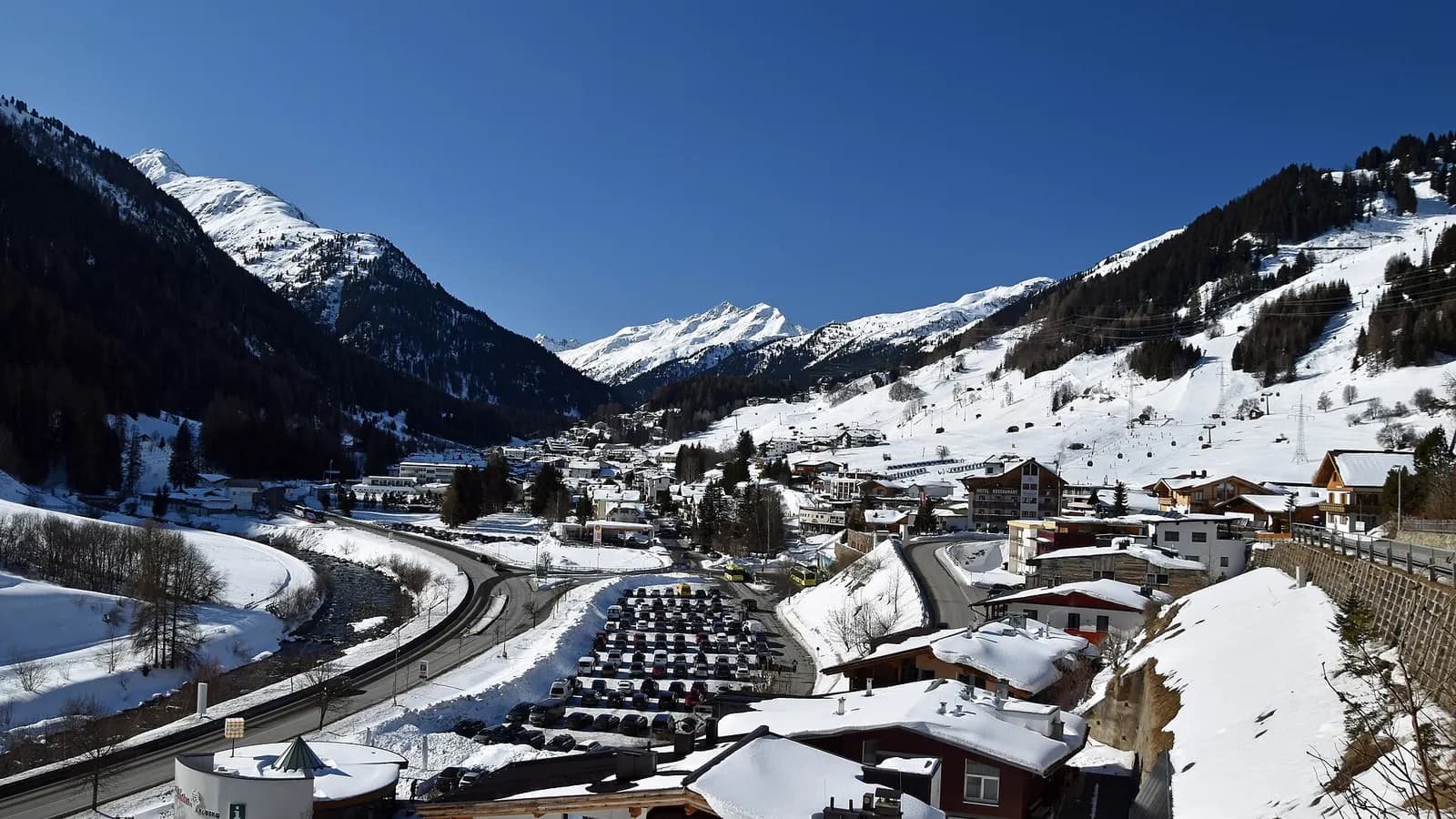 St. Anton am Arlberg im Winter, von oberhalb von Nasserein gesehen, mit Talstraße, Parkplatz und verschneiten Gipfeln unter blauem Himmel.