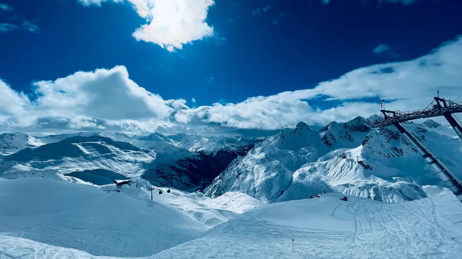 Verschneite Pisten und Gipfel in St. Anton am Arlberg unter tiefblauem Himmel, rechts ein Liftmast, in der Ferne eine Gondelbahn.