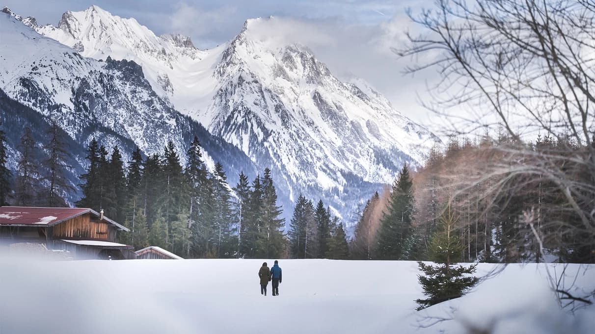 Twee wandelaars steken een besneeuwde weide over richting de bergen van de Arlberg, langs een houten berghut.