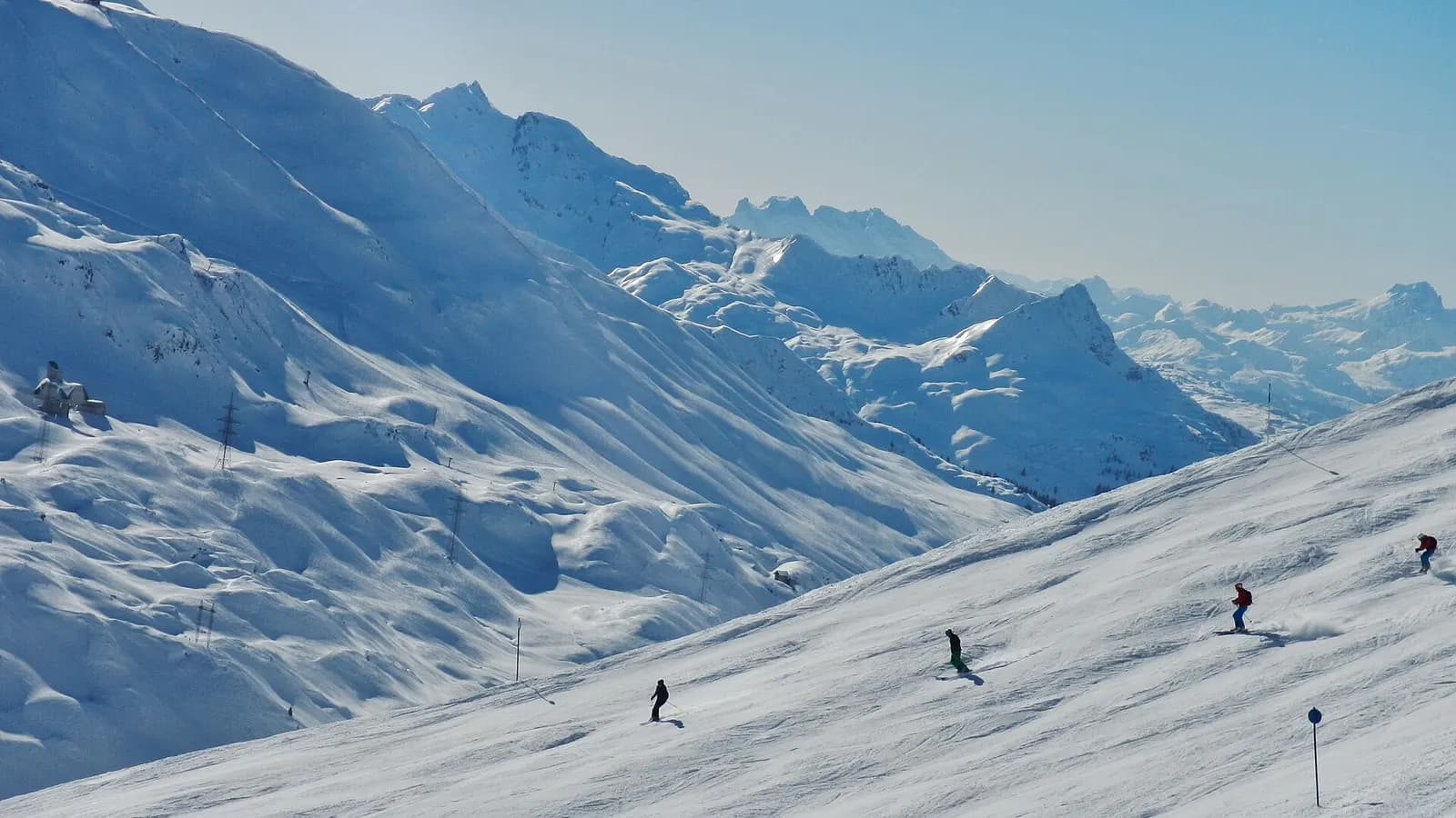 Vier skiërs op een brede, zonnige piste boven St. Anton am Arlberg, met besneeuwde bergen richting St. Christoph op de achtergrond.